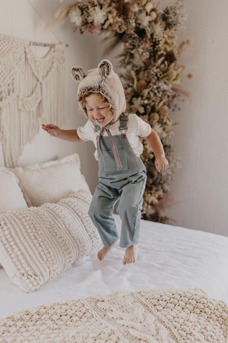 May include: A child wearing a brown and white faux fur bear hat jumps on a white bed with a cream-colored knitted throw blanket. The child is wearing a white t-shirt and blue denim overalls.