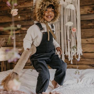 May include: A child wearing a brown furry animal hat and dark blue overalls is jumping on a bed with a pink blanket. The child is smiling and holding a brown furry tail.