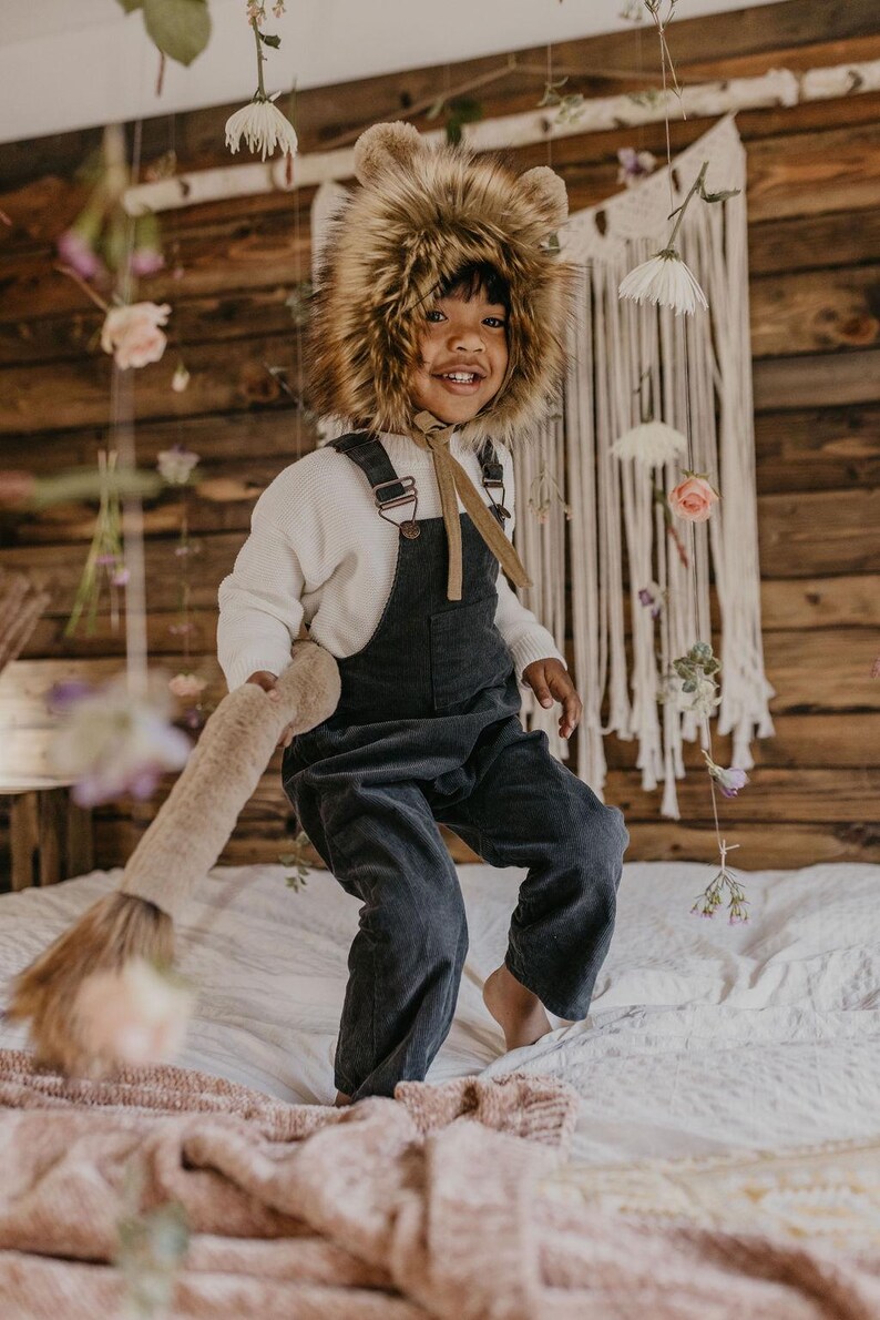 May include: A child wearing a brown furry animal hat and dark blue overalls is jumping on a bed with a pink blanket. The child is smiling and holding a brown furry tail.