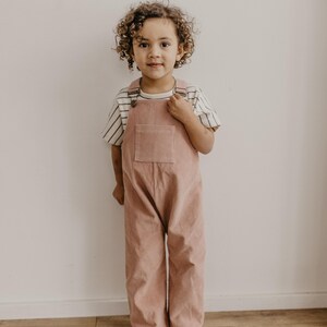 May include: A young child wearing a pink corduroy overall with a striped shirt. The child is standing on a wooden floor in front of a white wall.