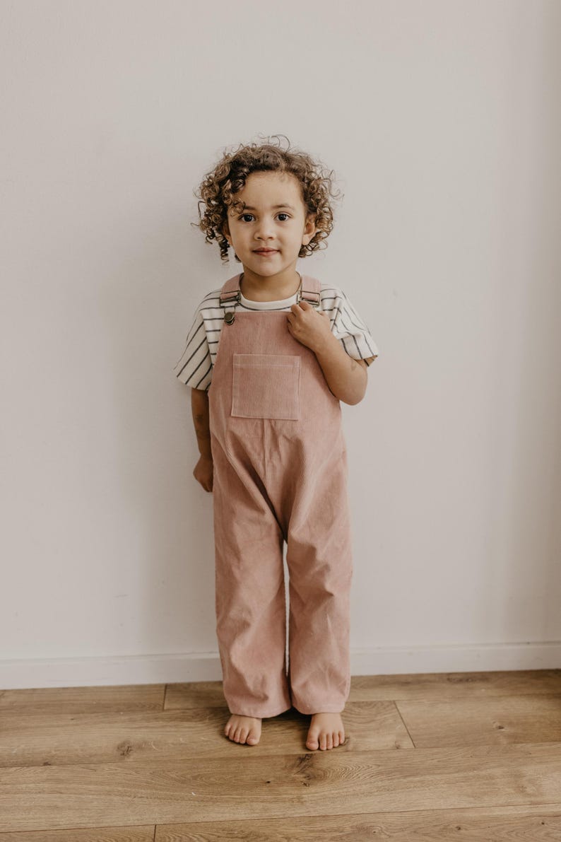 May include: A young child wearing a pink corduroy overall with a striped shirt. The child is standing on a wooden floor in front of a white wall.