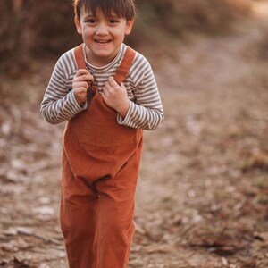 May include: A young boy wearing a striped long-sleeve shirt and brown overalls smiles while standing on a path in a wooded area.