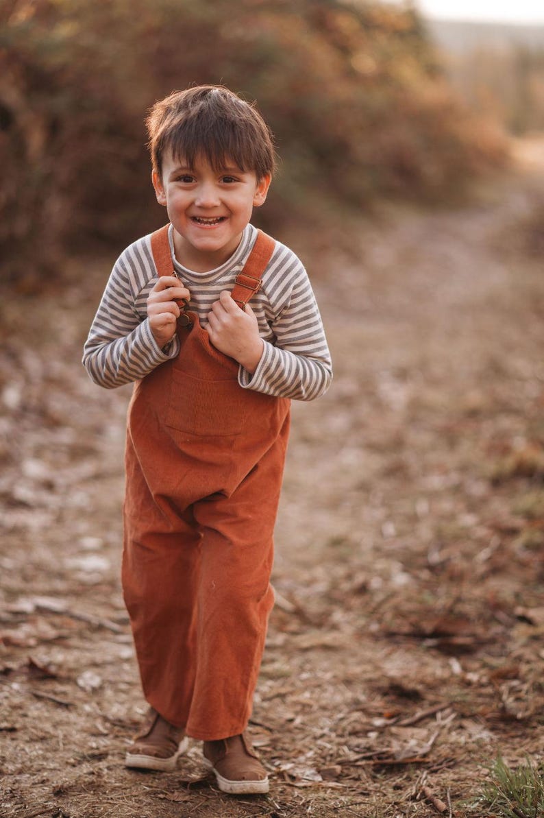May include: A young boy wearing a striped long-sleeve shirt and brown overalls smiles while standing on a path in a wooded area.