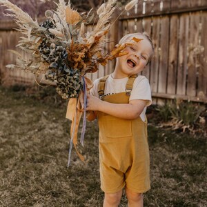May include: A young child wearing a yellow overall and a white t-shirt holds a bouquet of dried flowers. The bouquet includes dried grasses, pampas grass, and blue hydrangeas. The child is smiling and looking at the camera.