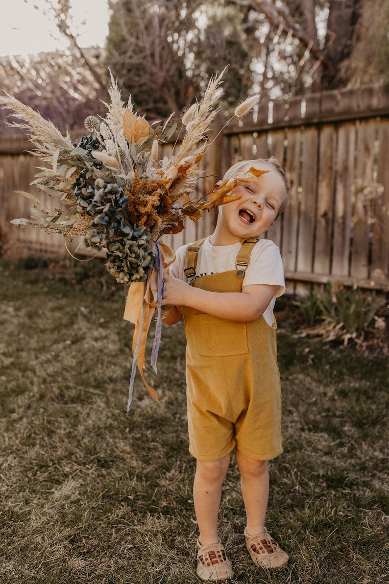 May include: A young child wearing a yellow overall and a white t-shirt holds a bouquet of dried flowers. The bouquet includes dried grasses, pampas grass, and blue hydrangeas. The child is smiling and looking at the camera.