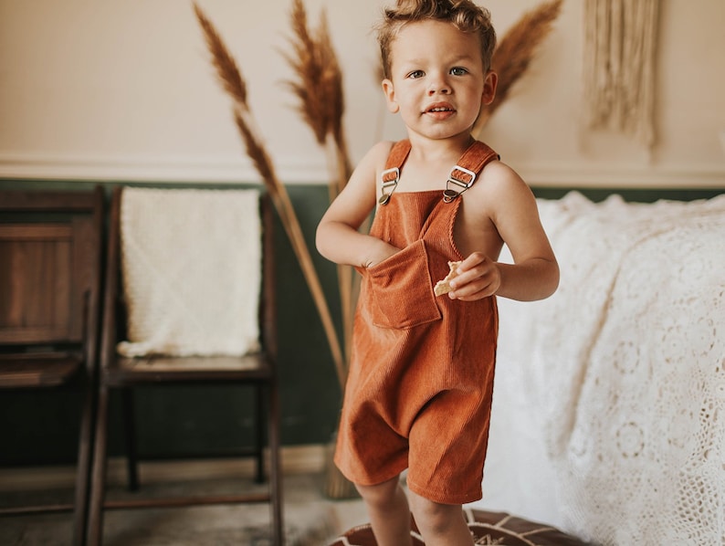 May include: A young child wearing a rust-colored corduroy overall with brown straps and a single front pocket. The child is standing in a room with a white blanket and wooden chairs in the background.