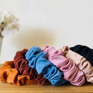 May include: Six fabric scrunchies in various colors, including brown, blue, pink, beige, and black. The scrunchies are arranged in a row on a wooden surface.