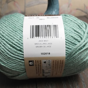 May include: A skein of Jade Mist yarn, a light green color, with a label that reads "Jade Mist, Brouillard Jade, Bruma de Jade" and a barcode. The label also includes the date 2021 06 18813, the code WL229329, and the number 102018.