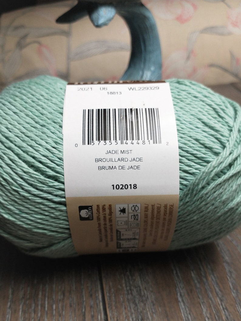 May include: A skein of Jade Mist yarn, a light green color, with a label that reads "Jade Mist, Brouillard Jade, Bruma de Jade" and a barcode. The label also includes the date 2021 06 18813, the code WL229329, and the number 102018.