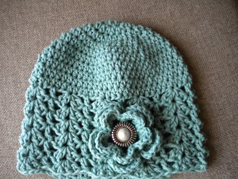 May include: A teal blue crocheted hat with a crocheted flower embellishment and a silver button in the center.