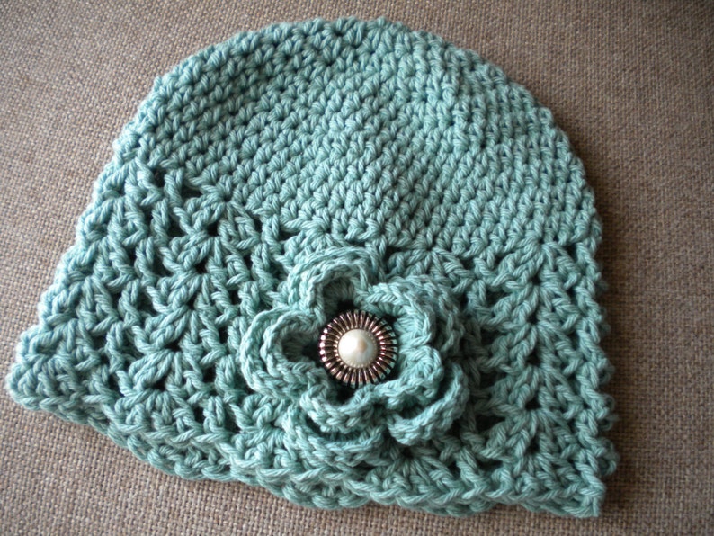 May include: A crocheted hat in a light blue color with a crocheted flower embellishment. The flower has a silver button in the center.