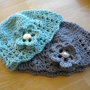 May include: Two crocheted hats with flower embellishments. The top hat is light blue with a white button, and the bottom hat is gray with a silver button.