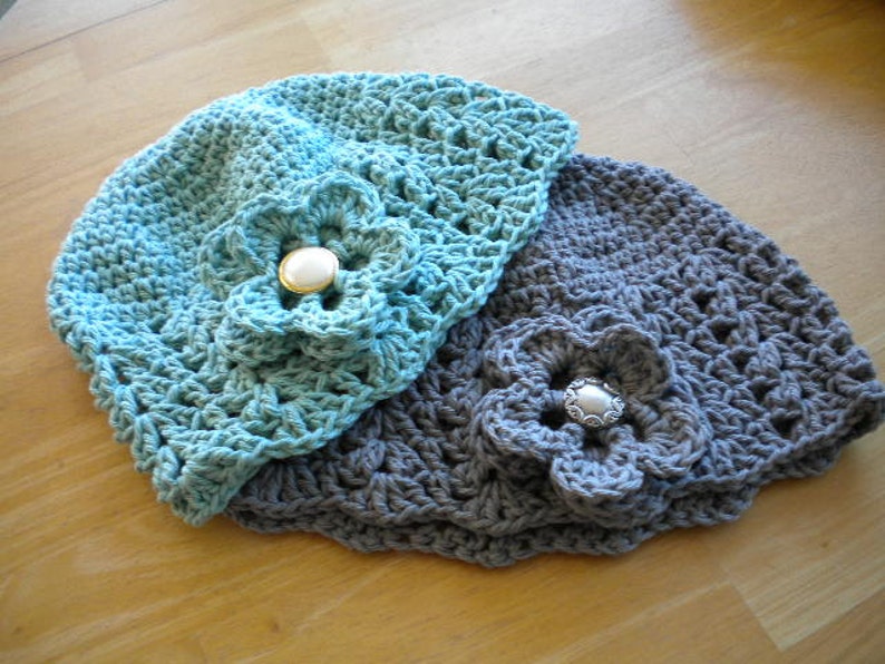 May include: Two crocheted hats with flower embellishments. The top hat is light blue with a white button, and the bottom hat is gray with a silver button.