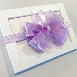 May include: A lavender satin and sheer ribbon bow headband. The headband is displayed on a white frame.