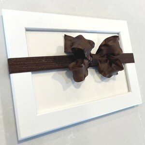 May include: A white picture frame with a brown ribbon tied in a bow in the center. The ribbon is attached to the frame with a brown elastic band.
