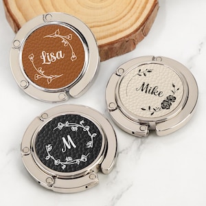 Puede incluir: Tres colgadores de bolso plateados con inserciones de cuero personalizadas. Uno tiene una inserción marrón con el nombre "Lisa", otro tiene una inserción crema con "Mike", y el tercero tiene una inserción negra con la letra "M". Cada uno está decorado con diseños florales.