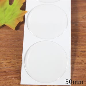 May include: A white rectangular sheet containing multiple clear, circular adhesive stickers. Each sticker has a diameter of 50mm, as indicated by the text printed in the bottom right corner of the sheet. The stickers are smooth and transparent.