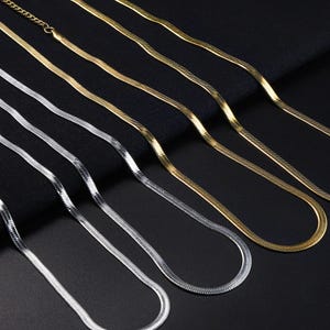 May include: A collection of gold and silver herringbone chain necklaces. The necklaces are displayed on a black surface, showcasing their sleek, flat, and flexible design. The chains vary in color, with some appearing gold and others silver.