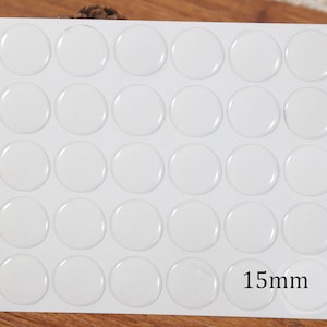 May include: A white sheet containing twenty-five clear, circular, adhesive-backed stickers arranged in a five-by-five grid. Each individual sticker has a smooth, domed surface and a diameter of 15mm as indicated by the text on the bottom right.