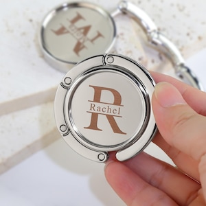 Puede incluir: Un colgador de bolsos circular de metal plateado con acabado pulido e incrustación central. La incrustación muestra una letra R grande de color marrón con el nombre Rachel impreso en una línea horizontal en el centro de la letra en fuente serif.
