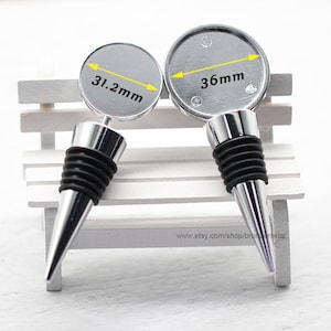 Könnte beinhalten: Zwei silberne Flaschenverschlüsse mit schwarzen Gummidichtungen und konischen Spitzen. Die runden Oberteile haben einen Durchmesser von 31,2 mm und 36 mm. Die Verschlüsse werden auf einer weißen Bank präsentiert.