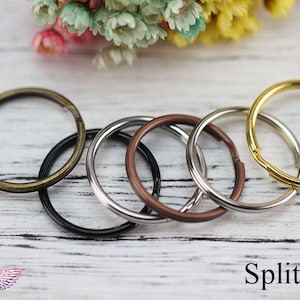 Puede incluir: Cinco anillos partidos de diferentes tamaños y colores, incluyendo oro, plata, negro y bronce. Los anillos están dispuestos en un círculo sobre una superficie de madera blanca.