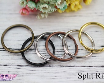 May include: Five split rings in different sizes and colors, including gold, silver, black, and bronze. The rings are arranged in a circle on a white wooden surface.