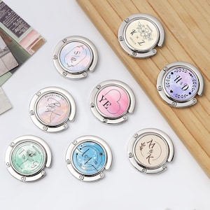 Puede incluir: Una colección de colgadores de bolso plateados personalizados con varios diseños. Cada colgador presenta un diseño circular con iniciales, fechas y detalles florales o celestiales. Los colores incluyen rosa, azul y crema, con un fondo de madera.