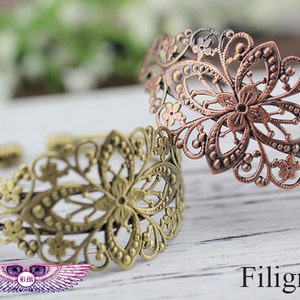 Puede incluir: Dos pulseras de filigrana, una en oro y otra en cobre, con intrincados diseños florales. Las pulseras están sobre una superficie de madera blanca.