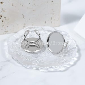 May include: Two silver ring blanks with round, flat, silver bases. The bases are about 2.5 cm in diameter. The ring blanks are on a white, textured surface.