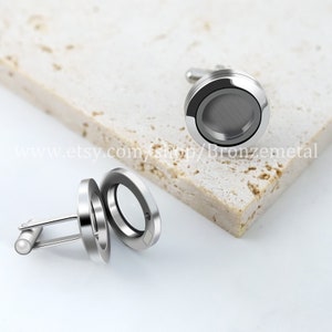 May include: Silver-toned cufflinks with a circular design. One cufflink is open, revealing a clear interior, while the other is closed, displaying a brushed metal surface. The cufflinks are set against a light-colored, textured background.