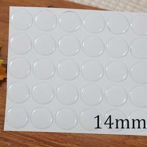 Puede incluir: Una hoja de 36 pegatinas redondas blancas adhesivas, cada una con un diámetro de 14 mm. Las pegatinas están dispuestas en una cuadrícula sobre un fondo blanco. El texto "14mm" está impreso en negro en la parte inferior de la hoja.