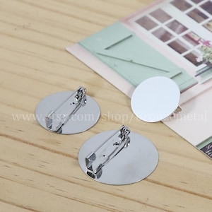 Puede incluir: Dos bases de broche redondas en blanco de metal plateado con cierre de pasador. Las bases son lisas y brillantes.