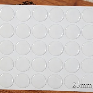 May include: A sheet of thirty clear, circular adhesive stickers arranged in a five-by-six grid. Each sticker has a smooth, domed surface and a diameter of 25mm, printed in the bottom right corner of the white backing sheet.