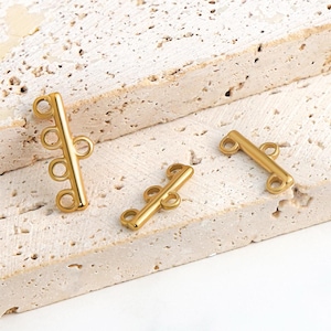 May include: Three gold-coloured jewellery clasps are displayed on a textured, light-coloured surface. The clasps have a bar shape with multiple loops for attaching jewellery pieces. The clasps are made of a shiny, metallic material.