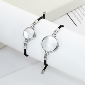 Peut inclure: Deux bracelets assortis. Chaque bracelet est orné d'un charm rond et transparent en forme de loupe, serti dans un cadre en métal argenté. Les charms sont attachés à un cordon noir. Les bracelets sont présentés sur une surface blanche.