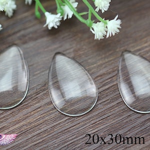 May include: Three clear glass teardrop-shaped cabochons, each measuring 20mm by 30mm, are arranged on a wooden surface.