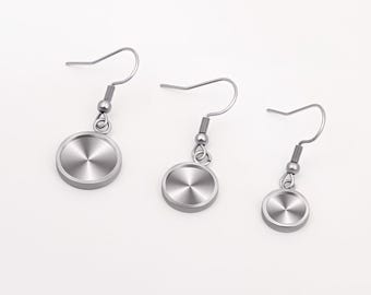 Può includere: Tre orecchini color argento con un design circolare. Ogni orecchino presenta un gancio, una piccola sfera, una molla e un pendente circolare con un motivo radiale strutturato. Sono presentati su sfondo bianco.