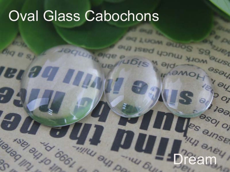 Pode incluir: Tr&ecirc;s caboch&otilde;es ovais de vidro transparente de tamanhos variados. O texto "Oval Glass Cabochons" est&aacute; no topo. A palavra "Dream" est&aacute; na parte inferior. Os caboch&otilde;es t&ecirc;m texto vis&iacute;vel atrav&eacute;s deles.