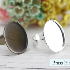 Op de afbeelding: Twee messing ringen met ovale glazen cabochons. Een ring heeft een zilverkleurig cabochon en de andere een bronskleurig cabochon. De ringen liggen op een wit houten oppervlak. Messing ring.