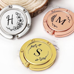 Peut inclure: Trois porte-sacs personnalisés en argent, or rose et or. Chacun présente une initiale différente : "H" avec des motifs floraux, "M" avec une couronne de laurier et "S" avec un motif floral. Ces cintres pliables sont conçus pour tenir des sacs à main.