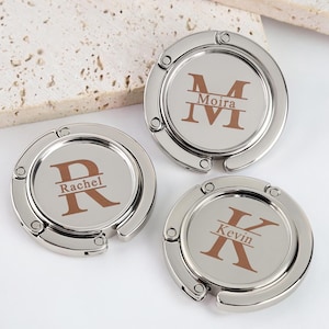 Puede incluir: Tres colgadores de bolsos circulares de metal plateado con un mecanismo de gancho plegable. Cada colgador presenta una incrustación central con una letra monograma marrón y un nombre impreso: M para Moira, R para Rachel y K para Kevin.