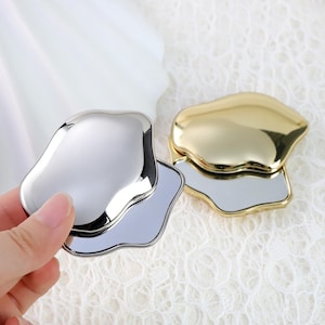 May include: Two compact mirrors with organic shapes. One is silver, held by a hand, and the other is gold. Both have a reflective surface and a matching base. The background is a white, textured fabric.