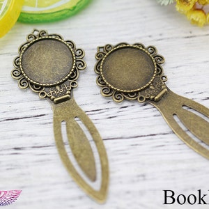 Puede incluir: Dos colgantes de marcapáginas de metal dorado antiguo con diseños de filigrana ornamentados e inserciones de cabujón de vidrio redondas. Los marcapáginas están sobre una superficie de madera blanca.