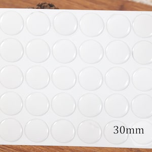 May include: A white sheet containing twenty-five clear, circular, adhesive-backed epoxy stickers arranged in a five-by-five grid. Each individual sticker has a smooth, domed surface and the text 30mm is printed on the bottom right corner.