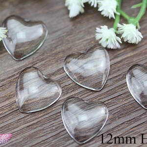 May include: Five clear glass heart-shaped beads, each 12mm in size, are arranged on a wooden surface. White floral accents are visible in the background. The beads are transparent, allowing the wood grain to show through.