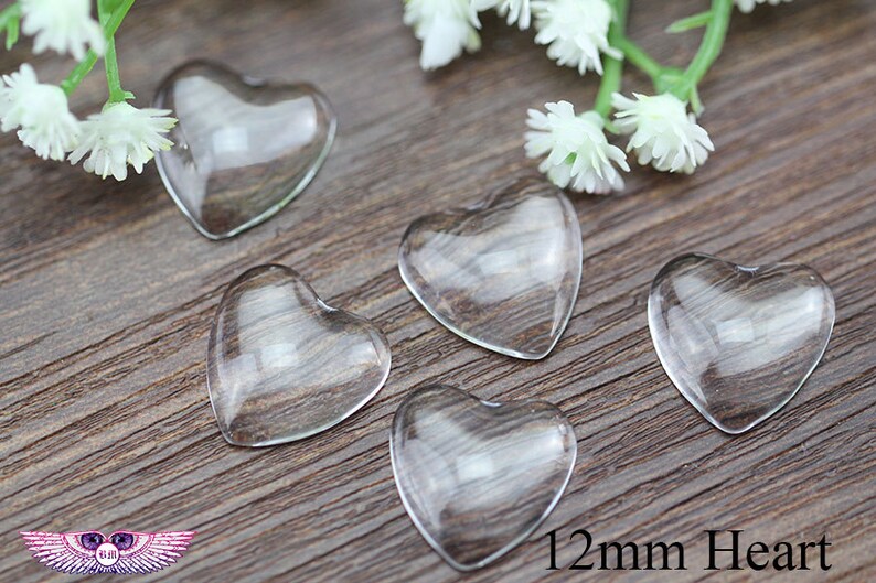May include: Five clear glass heart-shaped beads, each 12mm in size, are arranged on a wooden surface. White floral accents are visible in the background. The beads are transparent, allowing the wood grain to show through.