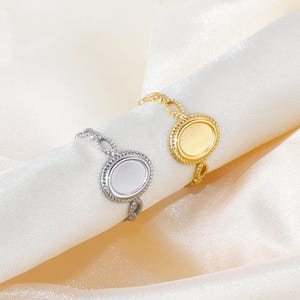 Puede incluir: Dos anillos, uno plateado y otro dorado, se muestran sobre una superficie de satén blanco. Cada anillo presenta una pieza central ovalada con un borde texturizado y un delicado diseño de cadena. El anillo dorado tiene un acabado pulido, mientras que el plateado tiene un acabado mate.
