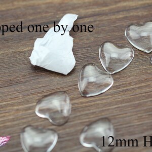 May include: Clear glass heart-shaped beads, each measuring 12mm, are scattered on a wooden surface. The text "Wrapped one by one" is visible. A small piece of white tissue paper is also present.