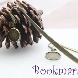 May include: A pair of bronze metal bookmarks with round, empty, metal charms. The bookmarks are laying on a white surface with a pine cone in the background. The text "Bookmarks" is written in purple below the bookmarks.